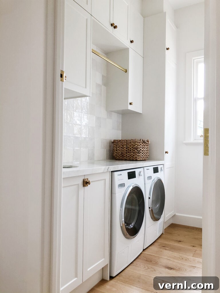 Why Homeowners Move Laundry Rooms Upstairs — And You Should Too 4 Bright and airy laundry room upstairs featuring white cabinetry with brass accents, a hanging rail, marble countertop, and side-by-side washer and dryer on natural wood flooring.