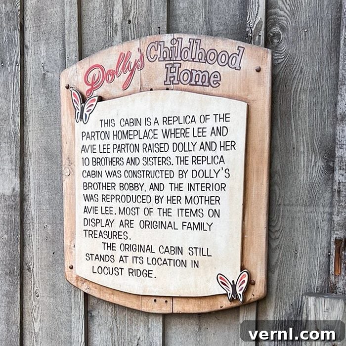 Wooden sign reads: “Dolly’s Childhood Home. This cabin is a replica of the Parton home where Dolly Parton and her family lived, featuring original family treasures. The original still stands in Locust Ridge.”.