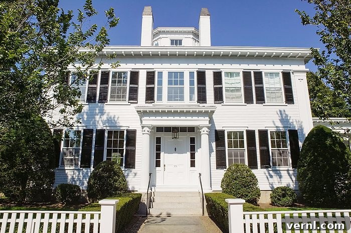 Why New Houses Look Alike and Why They're All White 2 A large white two-story house with black shutters, a central front door, symmetrical windows, two chimneys, and a white picket fence, surrounded by green trees and shrubs, under a clear blue sky.